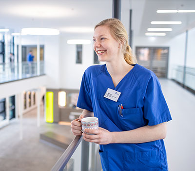 Auszubildende Gesundheits- und Krankenpflegerin im Flur eines Krankenhauses, lehnt am Treppengeländer mit einer Kaffeetasse in der Hand, Ausbildung in der Region, Niedersachsen