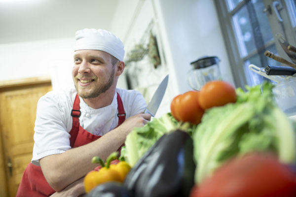 Auszubildender Koch Leonardo in der Kueche mit einem Messer, im Vordergrund Auberginen, Salat, Tomaten, Paprika