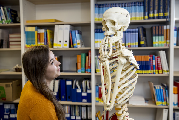 Auszubildende Gesundheits- und Krankenpflegerin Lea neben einem Skelett, vor einem Regal mit Büchern und Akten
