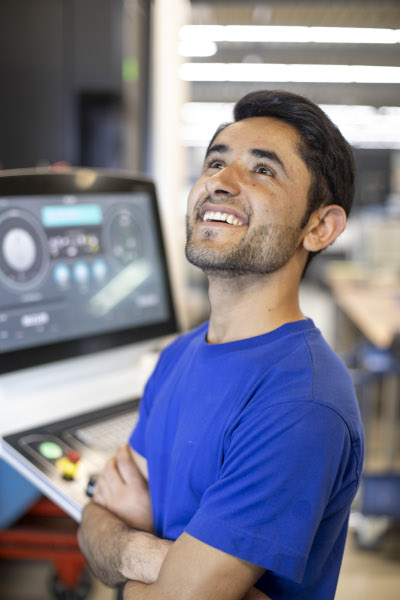 Auszubildender Zerspanungsmechaniker Obaidulah mit verschraenkten Armen vor einer technischen Anlage